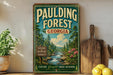 A vintage-style metal sign for Pauling Forest in Georgia, featuring a scenic illustration of a forest and a river, is displayed on a shelf next to a wooden cutting board and some potted plants.
