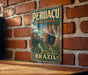A vintage-style metal sign displaying the name "PERUÇA" and "RIVER CANYON" in large letters, along with a scenic illustration of a river and mountains, is mounted on a brick wall.