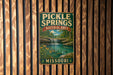 A vintage metal sign for Pickle Springs Natural Area in Missouri, featuring a scenic landscape with a waterfall and flowers.