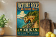 A vintage-style metal sign advertising the Pictured Rocks Lakeshore Trail in Michigan, featuring a scenic image of a cliff overlooking a lake.