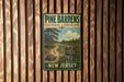 A vintage-style metal sign for Pine Barrens Cultural Landscape in New Jersey, featuring a scenic illustration of a forest and a winding path.