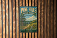 A vintage metal sign for Pine Hills Nature Preserve in Indiana, featuring a scenic landscape with a winding path and pine trees.