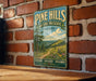 A vintage-style metal sign for Pine Hills Nature Preserve in Indiana, featuring a scenic landscape with a winding path and wildflowers.
