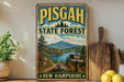A vintage-style metal sign for Pisgah State Forest in New Hampshire, featuring a scenic landscape with a mountain, river, and pine trees.