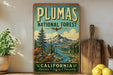 A vintage-style sign for the Plumas National Forest in California, featuring a scenic mountain landscape with pine trees and a river.