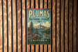 A vintage metal sign for the Plumas National Forest in California, featuring a scenic mountain landscape with pine trees and a river.