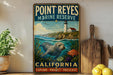 A vintage-style metal sign for Point Reyes Marine Reserve in California, featuring a lighthouse, seal, and marine life, is displayed on a shelf next to a wooden cutting board and a potted plant.