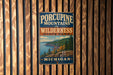 A vintage-style metal sign for Porcupine Mountains Wilderness State Park in Michigan, featuring a scenic landscape with mountains, forests, and a lake.