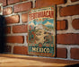 A vintage metal sign with a colorful illustration of Teotihuacan, the ancient city in Mexico, is displayed on a brick wall.