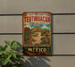 A vintage-style metal sign with a colorful illustration of Teotihuacan, the ancient city in Mexico, is displayed on a wall.