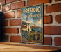 A vintage-style metal sign with the text "PRESIDIO OF SAN FRANCISCO, CALIFORNIA" and a graphic of a building and bridge, mounted on a brick wall.