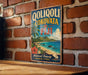 A vintage-style sign for Fiji hangs on a brick wall, featuring a tropical beach scene with palm trees, a blue ocean, and a white sailboat.