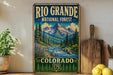A vintage-style sign for the Rio Grande National Forest in Colorado, featuring a mountain landscape with pine trees and a river flowing through it.