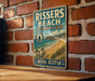 A vintage-style metal sign for Rissers Beach, a coastal park in Nova Scotia, Canada.