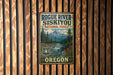 A vintage-style metal sign for Rogue River-Siskiyou National Forest in Oregon, featuring a scenic mountain landscape and a river.
