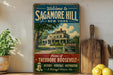 A vintage-style sign welcoming visitors to Sagamore Hill, New York, featuring a large house and a tree, with a wooden cutting board and lemons in the foreground.