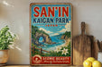 a vintage-style sign for San'In Kaigan Park, Japan, with a scenic landscape depicting a rocky arch, a beach, and a body of water. The sign is displayed on a shelf next to a wooden cutting board and a potted plant, creating a rustic and charming atmosphere.