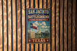 a vintage-style metal sign with the text "SAN JACINTO BATTLEGROUND" and "STATE HISTORIC SITE" displayed prominently. The sign also includes a depiction of a cannon and a Texas state monument, suggesting a connection to the state's history and military heritage.