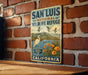 A vintage-style metal sign for the San Luis National Wildlife Refuge in California, featuring a bird and flowers, mounted on a brick wall.