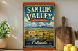 a vintage-style sign for San Luis Valley, Colorado, with a scenic landscape of mountains, a river, and a small building.