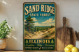 A vintage-style metal sign for the Sand Ridge State Forest in Illinois, featuring a scenic landscape with pine trees, sand dunes, and a lake.