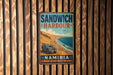 A vintage-style metal sign with the text "SANDWICH HARBOUR" and "NAMIBIA" is displayed on a wooden wall.