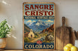 A vintage-style sign for the Sangre de Cristo National Heritage Area in Colorado, featuring a mountain landscape with a church and other buildings.