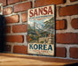 A vintage metal sign with the text "SANSA BUDDHIST MOUNTAIN MONASTERIES KOREA SOUTH KOREA" is displayed on a brick wall.