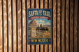 A vintage-style metal sign for the Santa Fe Trail in New Mexico, featuring a wagon on a dirt road with mountains in the background.