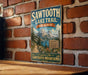 A vintage-style metal sign for the Sawtooth Lake Trail in Idaho, featuring a scenic mountain landscape and inviting visitors to explore the Sawtooth Mountains.