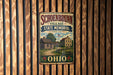 A vintage-style metal sign for Schoenbrunn Village State Memorial in Ohio, featuring a wooden house and a flag, mounted on a wooden wall.