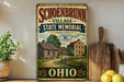 a vintage-style sign for Schoenbrunn Village, which is a State Memorial located in Ohio. The sign displays a picture of a log cabin and a small house, along with the text "Schoenbrunn Village State Memorial Since 1772" and "OHIO". The sign is placed on a shelf next to a wooden cutting board and a potted plant.