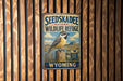 A vintage-style metal sign for the Seedskadee National Wildlife Refuge in Wyoming, featuring a bird perched on a wooden post against a backdrop of a blue sky and mountains.