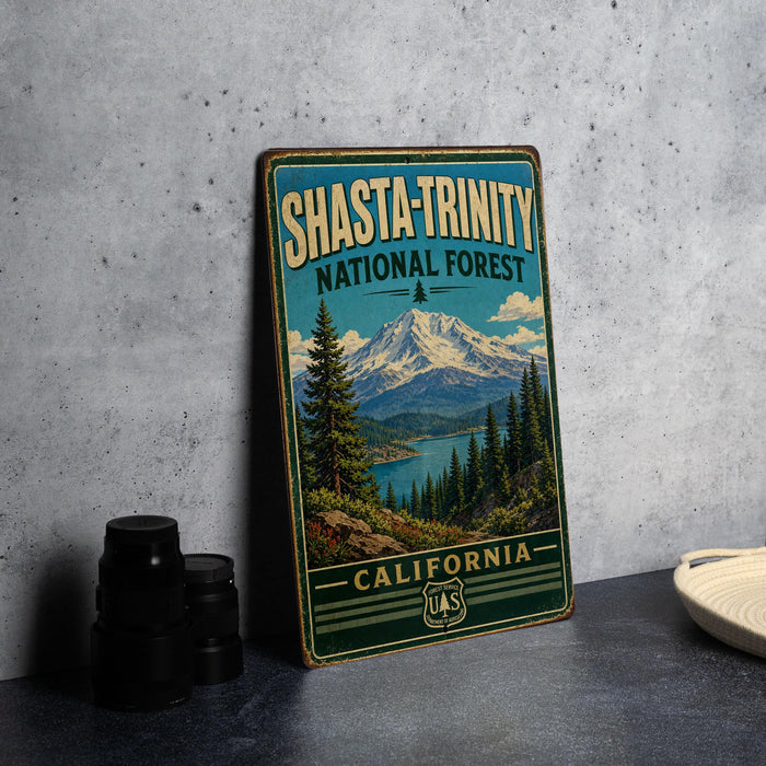 A vintage-style metal sign for Shasta-Trinity National Forest, featuring a mountain landscape and pine trees, is displayed on a gray countertop next to some black camera lenses.