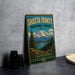 A vintage-style metal sign for Shasta-Trinity National Forest, featuring a mountain landscape and pine trees, is displayed on a gray countertop next to some black camera lenses.