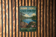 A vintage metal sign for Shawme-Crowell State Forest in Massachusetts, featuring a scenic landscape with a lake, pine trees, and a rocky outcrop.