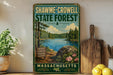 A vintage-style metal sign for Shawme-Crowell State Forest in Massachusetts, featuring a scenic landscape with pine trees, a lake, and a rocky outcrop.