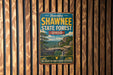 A vintage-style metal sign advertising Shawnee State Forest in Ohio, featuring a scenic landscape with a bridge and trees.