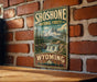 A vintage-style metal sign for Shoshone National Forest in Wyoming, featuring a mountain landscape with a waterfall.