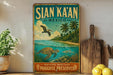 A vintage-style metal sign with a tropical beach scene, featuring a pelican, turtle, and palm trees, along with the text "SIAN KÁAN MEXICO" and "NATURAL HERITAGE PARADISE PRESERVED".