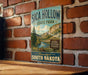 A vintage-style metal sign for Sica Hollow State Park, featuring a scenic landscape with a river and mountains, mounted on a brick wall.
