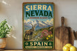 A vintage-style sign for Sierra Nevada Biosphere Reserve, featuring a mountain landscape with a goat and flowers, is displayed on a shelf next to a wooden cutting board and some lemons.