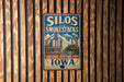A vintage-style metal sign with the text "Silos and Smoke stacks" and "National Heritage Area Iowa" is displayed on a wooden wall.