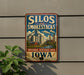 A vintage-style metal sign with the text "Silos and Smoke stacks" and "National Heritage Area Iowa" is displayed on a wall, with a plant visible to the left.