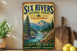 a vintage-style poster of the Six Rivers National Forest in California, with a scenic mountain landscape and a river flowing through it. The poster is displayed on a wall, and there are some potted plants and kitchen utensils, such as a knife and a spoon, visible in the foreground.