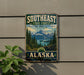 A vintage-style metal sign advertising the Southeast State Forest in Alaska, featuring a scenic mountain landscape with pine trees and a lake.