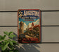 A vintage-style sign for St. Augustine, Florida, featuring a castle and other historic buildings, is displayed on a wall next to a plant.