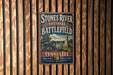 A vintage-style metal sign for the Stones River National Battlefield in Tennessee, featuring a cannon and a monument, mounted on a wooden wall.