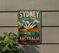 A vintage-style metal sign with the text "SYDNEY Opera House" and "AUSTRALIA Spectacular Harbour City" is displayed on a wall, with a plant to the left.