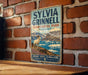 A vintage-style metal sign with the text "SYLVIA GRINNELL TERRITORIAL PARK NUNAVUT" and a depiction of a white ram standing on a rocky outcrop with a scenic background of mountains and water.